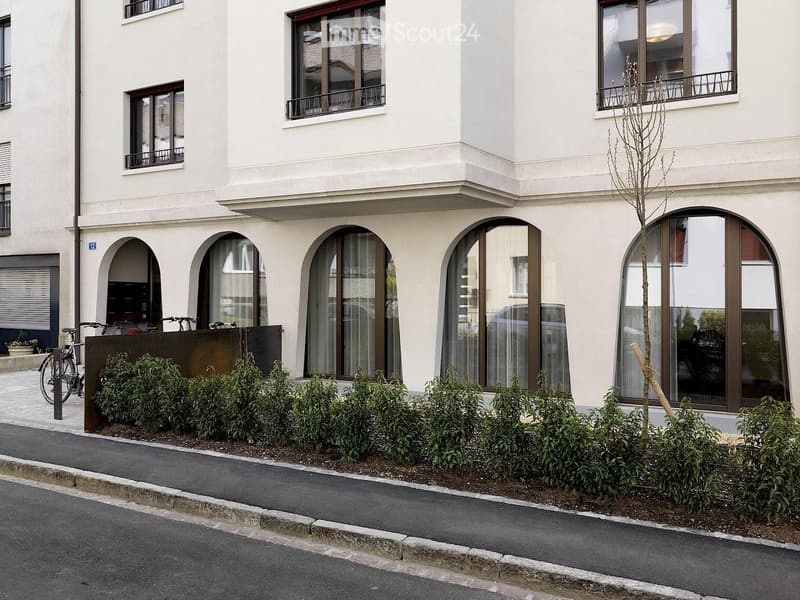 Modern white building with arched windows and balconies, surrounded by small shrubs and a bicycle.