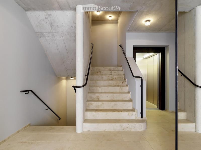Modern interior staircase with beige stone steps, black metal railings, and recessed lighting, leading to an open area with white walls and a raised ground floor.
