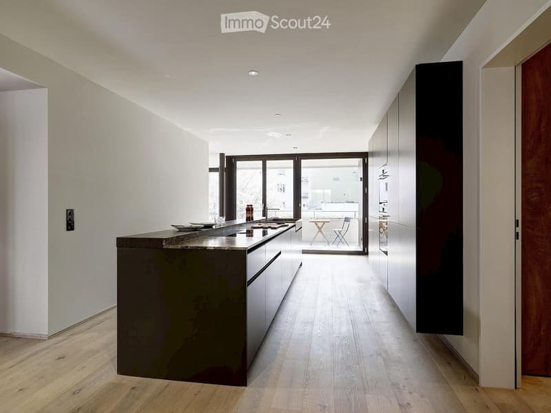 Modern kitchen with a dark countertop, white cabinets, and a sleek design. The space features a sink, a bottle on the counter, and a view to an outdoor area with a small table and chairs.