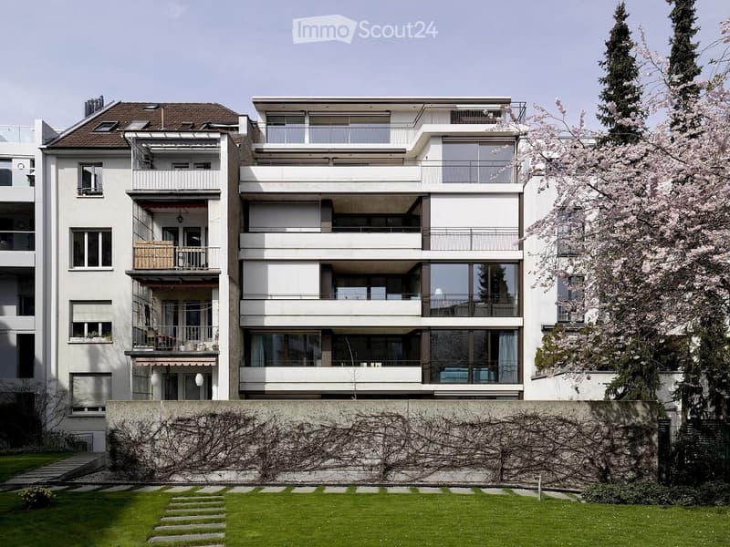 Modern multi-story residential building with white facade, large windows, balconies, and surrounding greenery including cherry blossom trees and a well-maintained lawn.