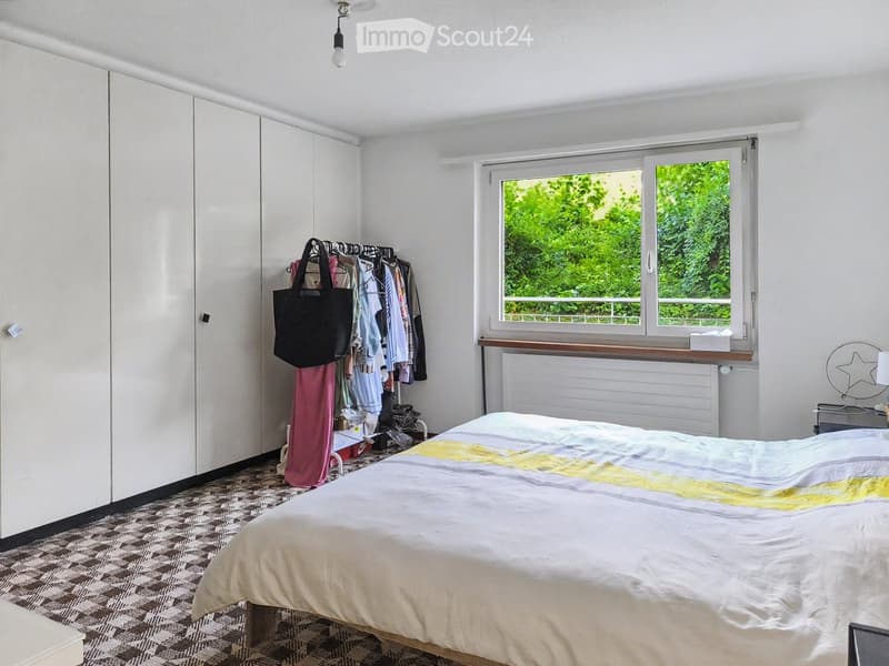 Bright bedroom with a large bed, white wardrobe, radiator, nightstand with lamp, and a clothing rack with clothes and a handbag.