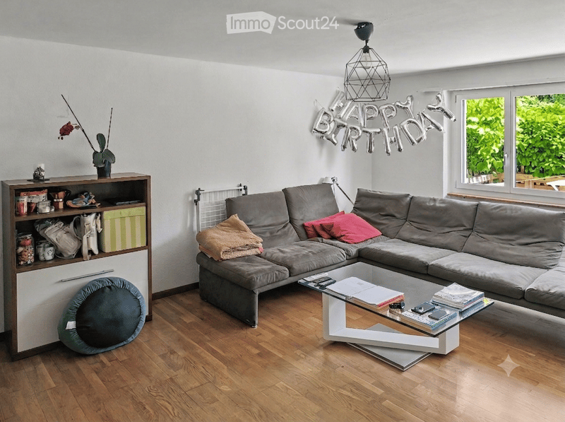 Modern living room with a grey sectional sofa, glass coffee table, bookshelf, and decorative pillows. The room features wooden flooring and large windows with a view of greenery outside.