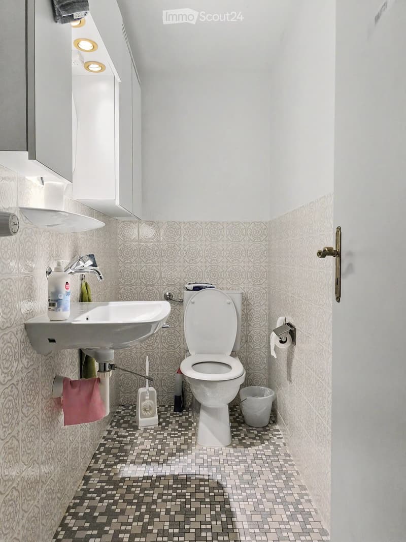 Modern bathroom with white walls, patterned tiles, a toilet, sink, and a small trash can. Includes a towel and a bottle on the sink.