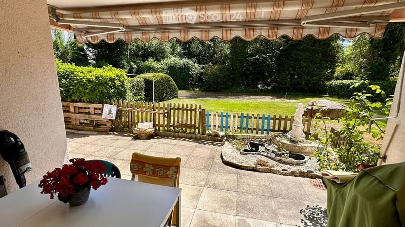 Outdoor patio with tiled floor, small table with red flowers, two chairs, and a small fountain area with a statue and surrounding greenery.