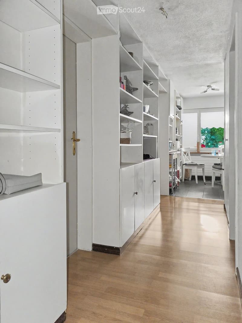 A modern, minimalist hallway with white walls, light wooden flooring, built-in white shelving units, and a small adjacent room with a table and chairs.