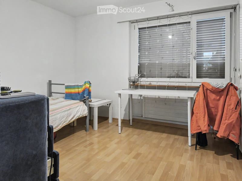 A small, well-lit bedroom with a bed, desk, and chair. The room features wooden flooring and white walls, with a window covered by blinds and string lights.