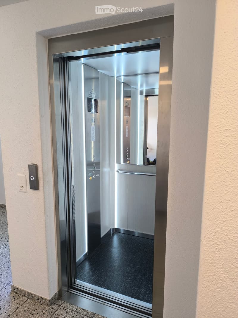 Modern elevator with glass doors, control panel, and illuminated interior, set in a hallway with light-colored walls and tiled flooring.