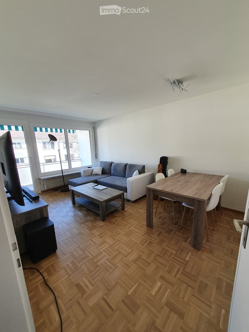 A modern living room with a grey sectional sofa, a wooden coffee table, a dining table with chairs, a television, and a floor lamp. The room features wooden herringbone flooring and large windows with blinds.