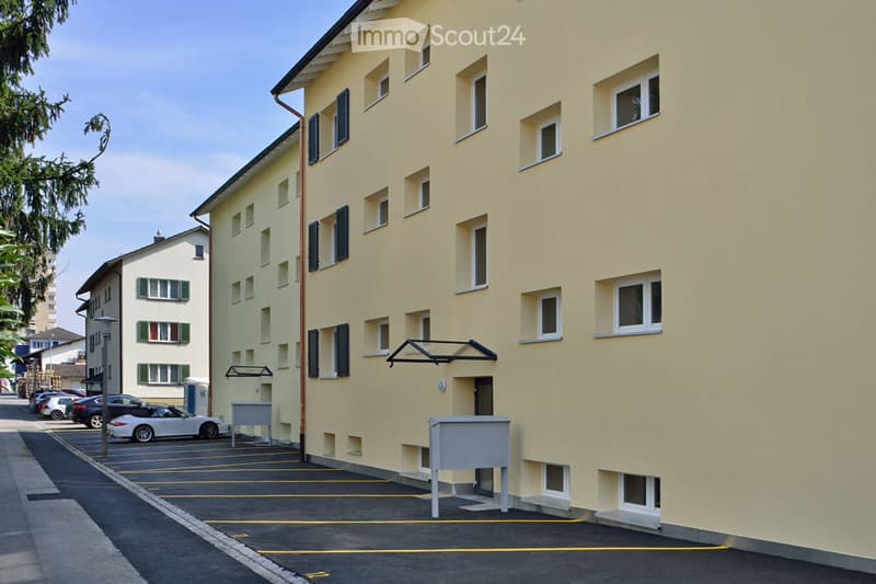 Multi-story residential building with light yellow facade, multiple windows, and a parking area in front.