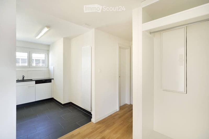 Bright, minimalist kitchen with white cabinets, a sink, a faucet, and a dishwasher. The space features a dark tiled floor in the kitchen area and wooden flooring in the adjacent living space.