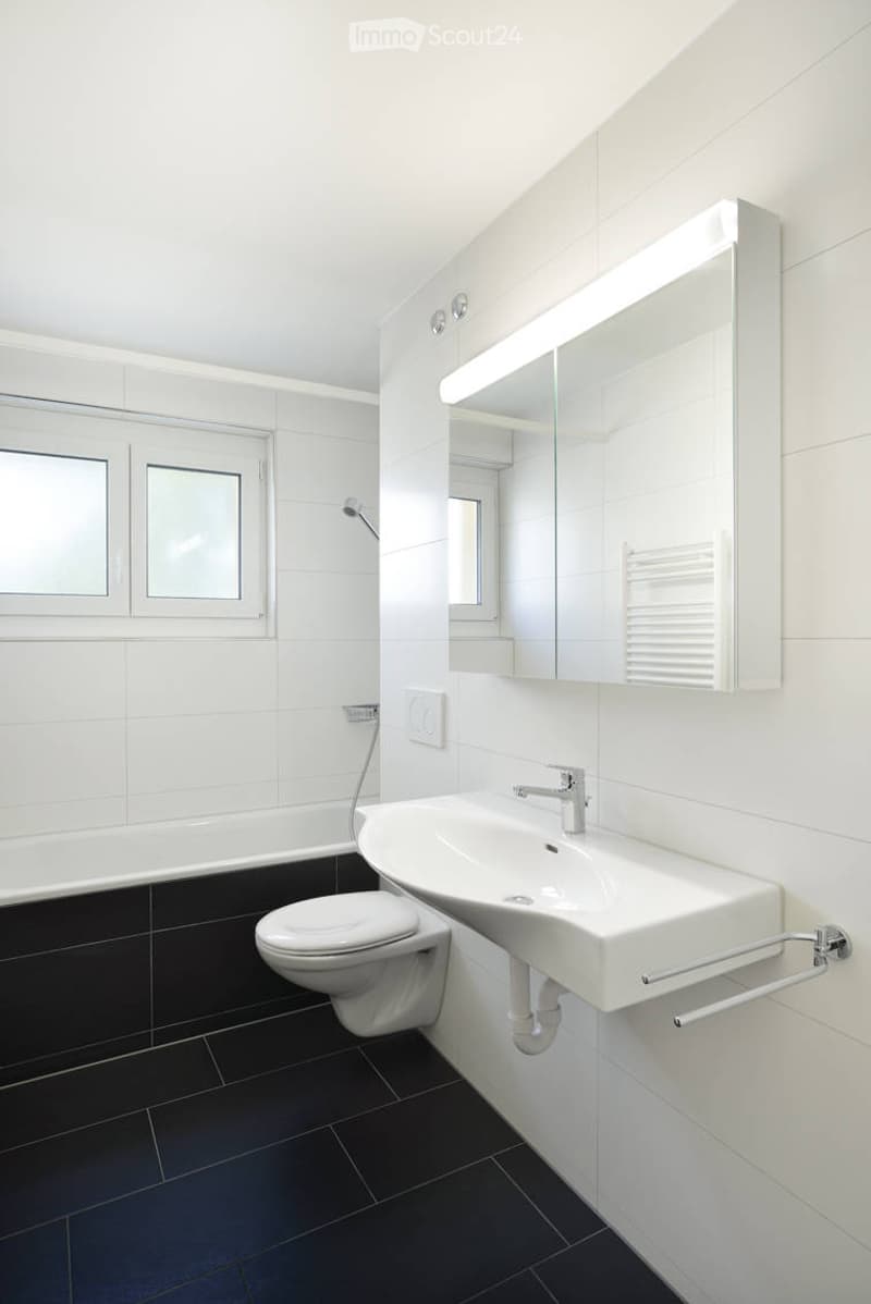 Modern bathroom with white tiles, black floor tiles, a white sink, toilet, bathtub, and a large mirror with built-in lighting.