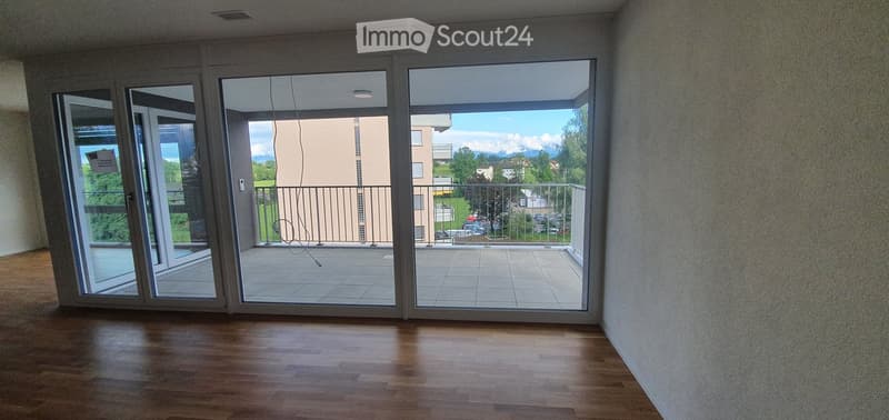 Empty room with wooden flooring, large sliding glass doors leading to a balcony with a metal railing, and a view of a building and greenery.
