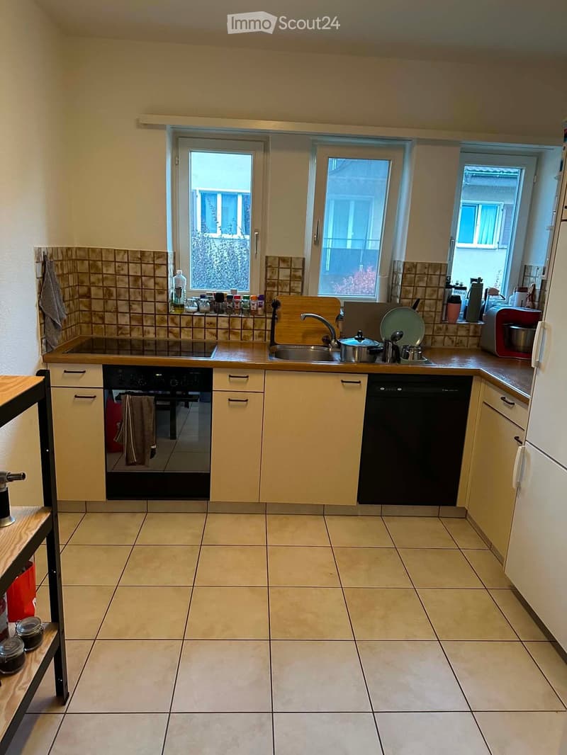 Bright kitchen with light-colored cabinets, tiled backsplash, and tiled floor. Features a sink, oven, dishwasher, and refrigerator, with various kitchen items on the counter and a small table to the left.