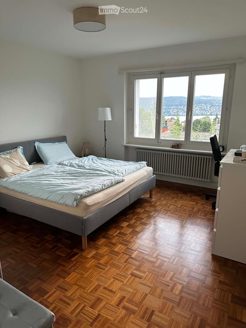 Bright bedroom with wooden flooring, large windows offering a scenic view, a neatly made bed with pillows and a blanket, a floor lamp, a chair, and a small desk.