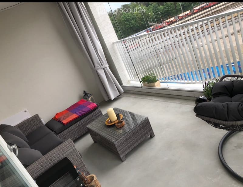 Balcony with a wicker sofa, coffee table, hanging chair, and a candle on the table. The balcony has a view of a train track and greenery.