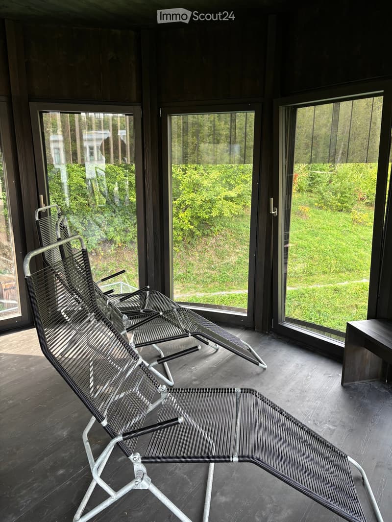 Modern balcony with large glass windows offering a view of greenery, featuring two metal lounge chairs and a small wooden table.