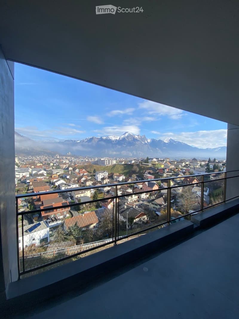Balcony with a modern glass railing, overlooking a town with houses and distant snow-capped mountains.