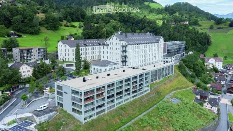 Aerial view of a large, multi-story building complex surrounded by greenery and smaller structures in a rural area.