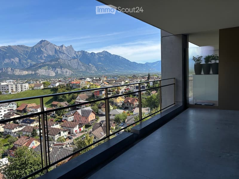 Balcony with a metal railing, overlooking a town with houses and mountains in the background.