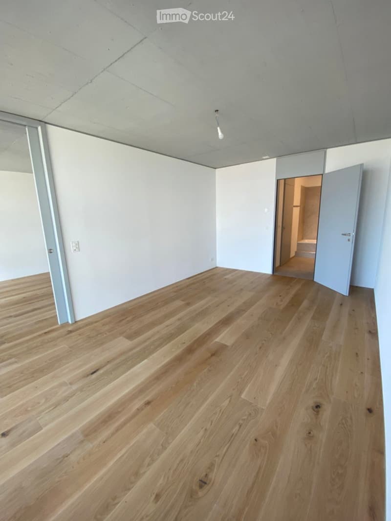 Empty room with light wooden flooring, white walls, and a visible doorway leading to another space.