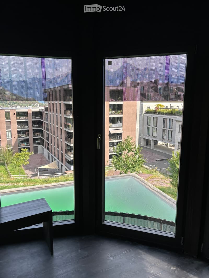 Modern room with large windows offering a view of a swimming pool, greenery, and surrounding buildings.