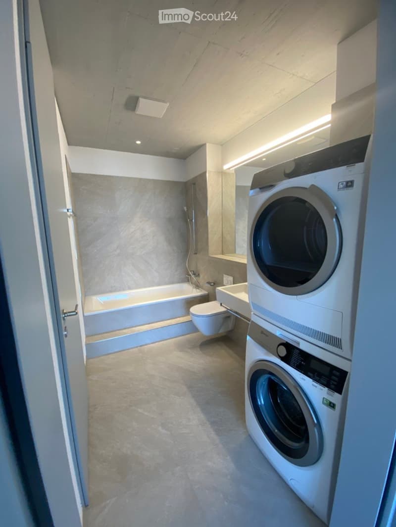 Modern bathroom with a bathtub, toilet, sink, and stacked washing machine/dryer unit. The space features light-colored tiles and a sleek design.