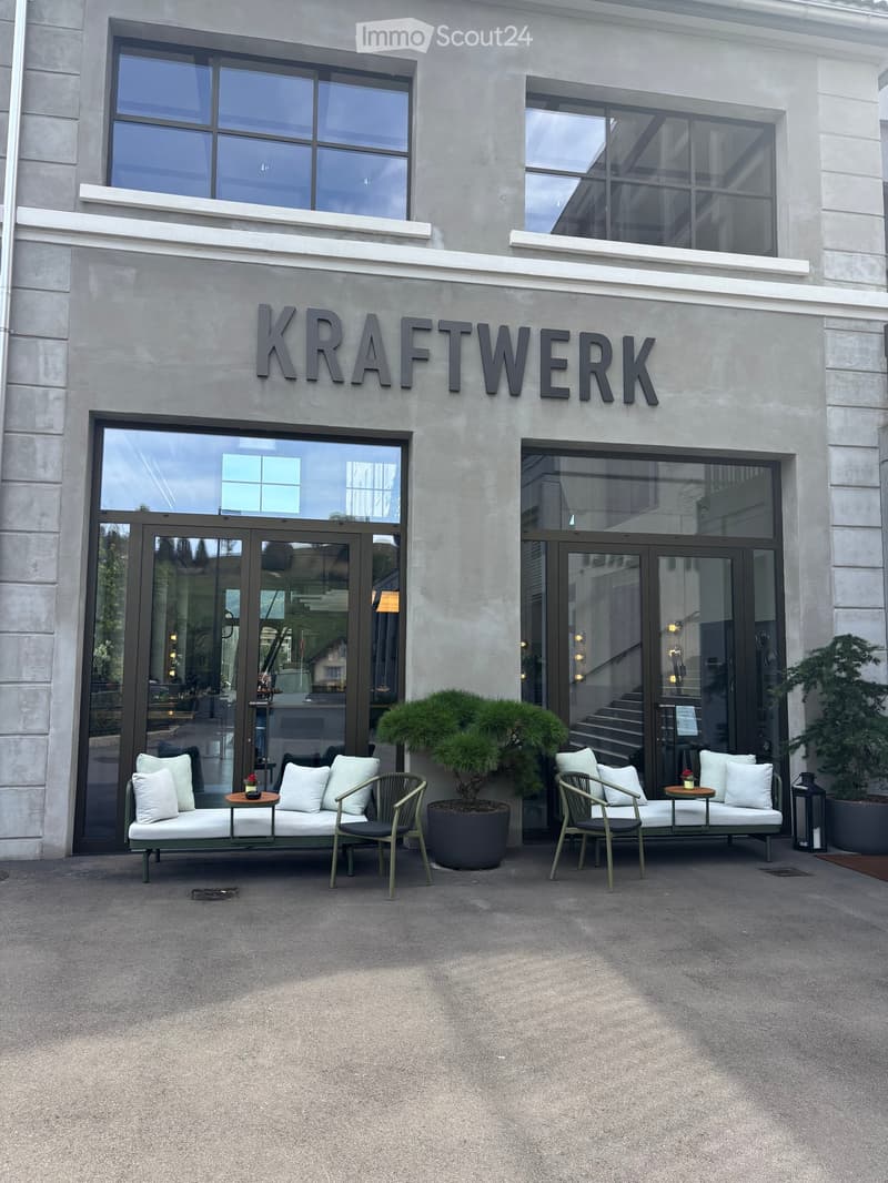 Modern outdoor seating area in front of a building with the sign "KRAFTWERK" above the entrance. Features two green sofas with white cushions, matching chairs, small tables, and potted plants.