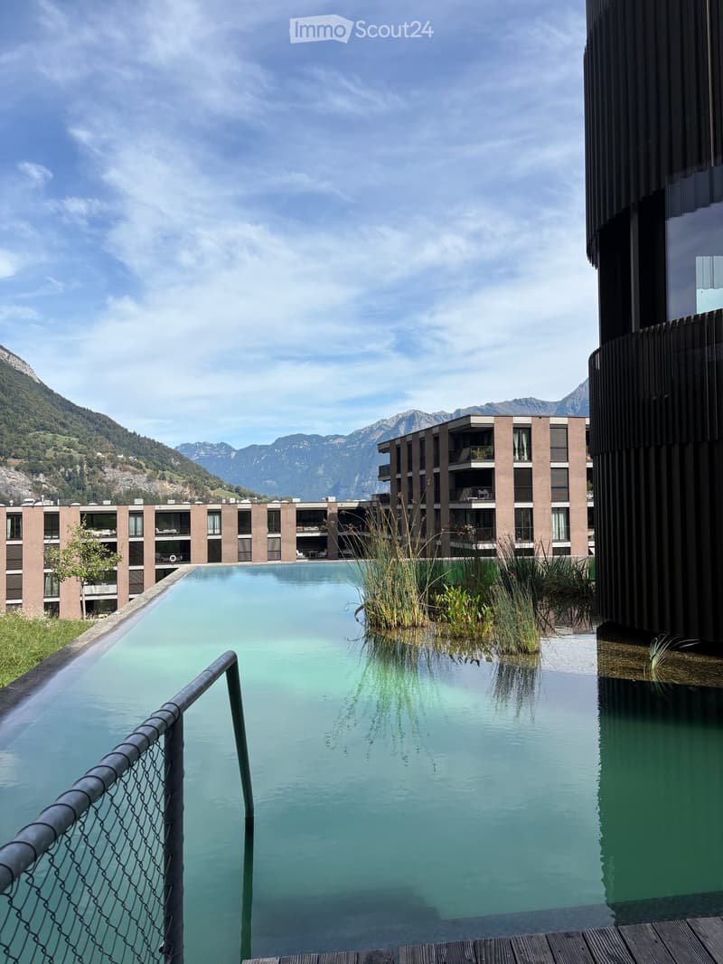 Outdoor area with a large swimming pool surrounded by modern buildings and a scenic mountain view.