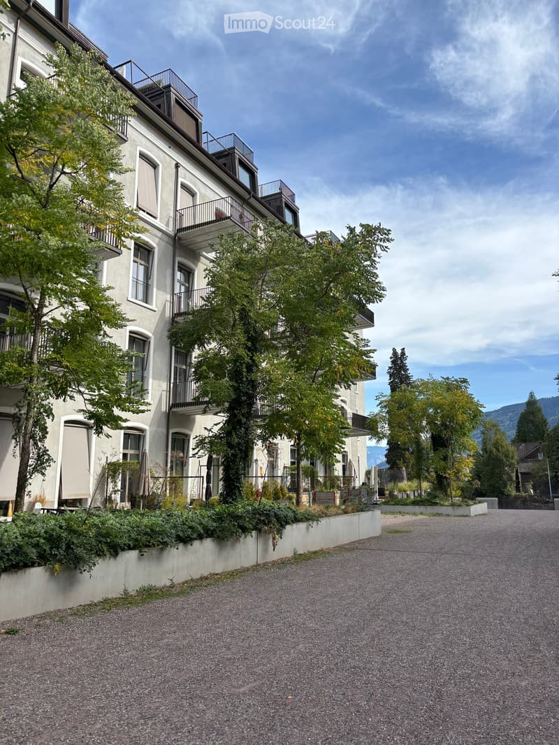 A multi-story residential building with balconies, surrounded by trees and greenery, with a gravel driveway in front.