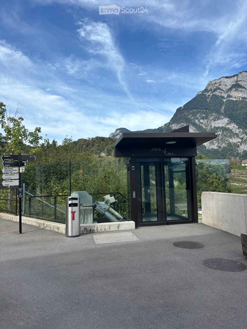 Modern building entrance with glass doors, signage reading "STOFFEL MELS," surrounded by greenery and a mountainous backdrop.