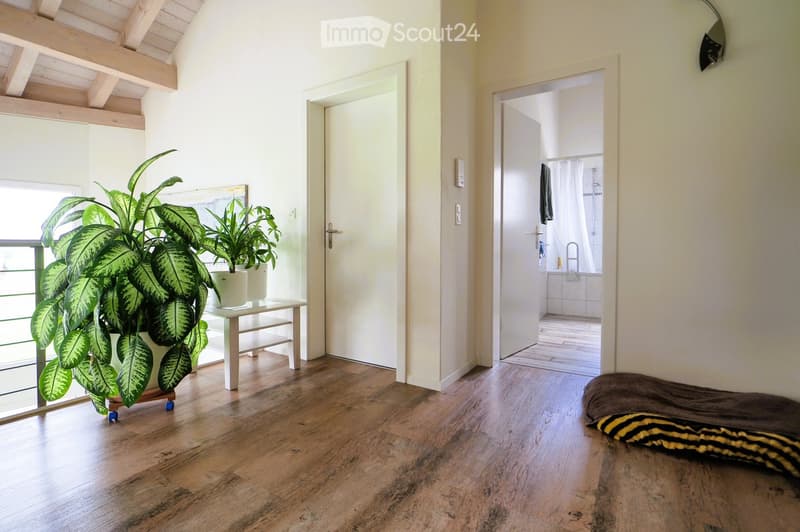 Bright hallway with wooden flooring, large window, small table with potted plants, and an open door leading to a bathroom.