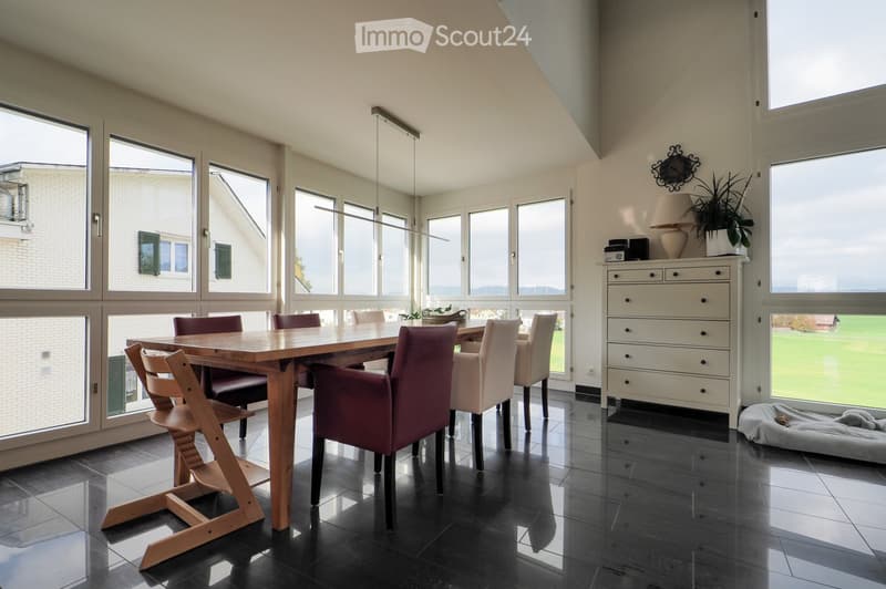Bright dining area with large windows, wooden dining table, chairs, white cabinet, lamp, plant, and dog bed.