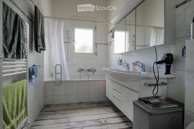 Modern bathroom with a bathtub, double sink, mirror, and wooden flooring. Includes towels, a hair dryer, and a small stool with a magazine.