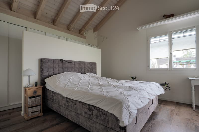 Modern bedroom with a large bed, grey headboard, white bedding, wooden nightstand with a lamp, and a large mirror on the wall. The room features wooden flooring and large windows with blinds.