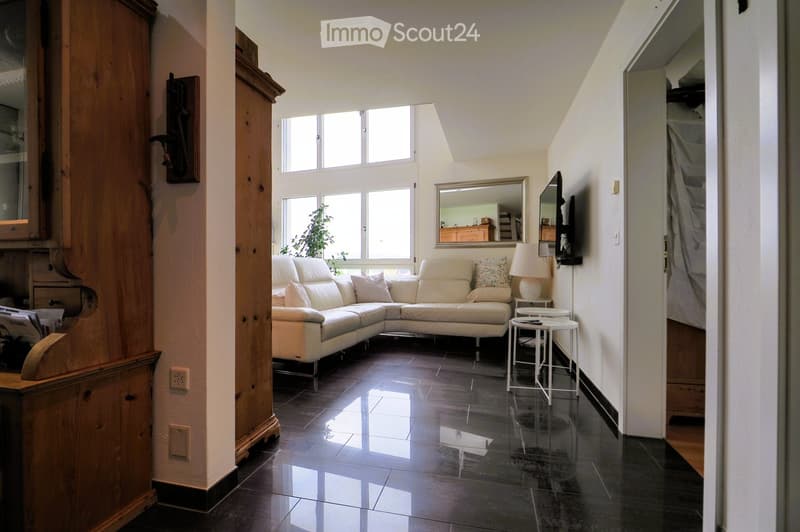 Bright living room with a white sectional sofa, small round tables, a lamp, and a large mirror. The room features large windows and a dark tiled floor.