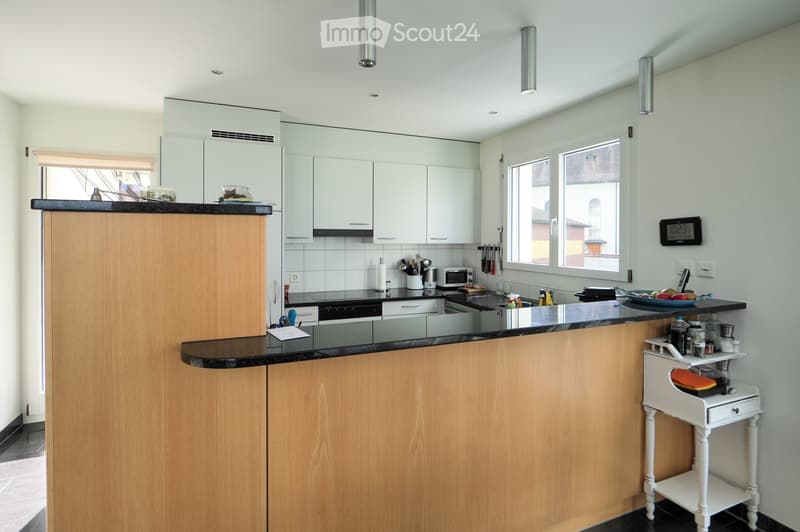 Modern kitchen with light wooden cabinets, black countertops, and stainless steel appliances. Features an island with a black countertop, various kitchen utensils, and a small white side table with items on it.
