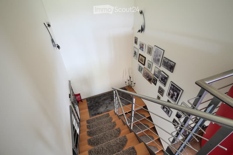 Spiral staircase with wooden steps, metal railing, and framed pictures on the wall. The staircase features a small landing area with a candle holder and a red bag on the floor.
