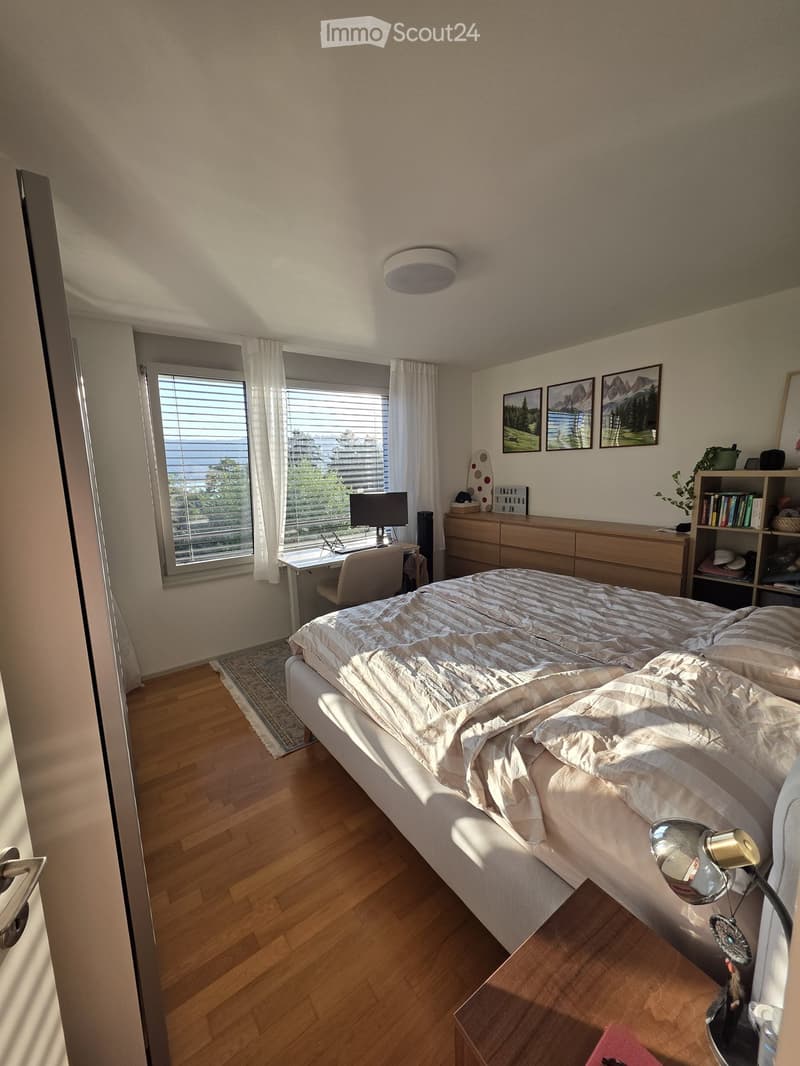 Modern bedroom with a large bed, nightstand, desk, and large window with blinds. The room features wooden flooring and several decorative items including picture frames and a shelf.