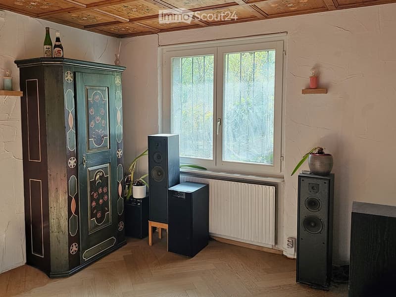 A cozy room with wooden flooring, a large window, a green wardrobe with floral designs, speakers, a small stool, and a radiator.