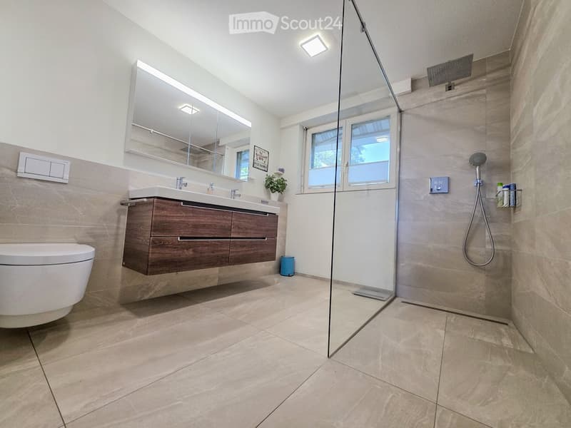 Modern bathroom with beige tiles, a wooden vanity with a sink, a toilet, a large mirror, and a glass-enclosed shower with a handheld showerhead.