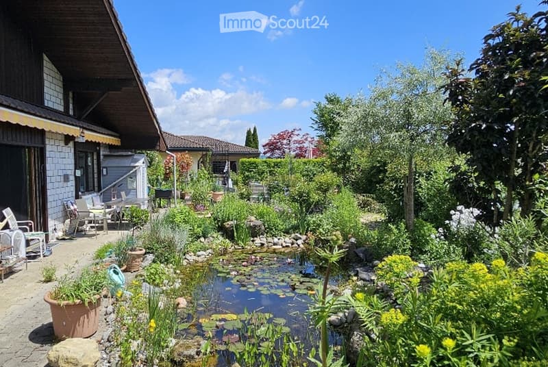 Garden with a pond, surrounded by lush greenery, flowers, and trees. Includes patio furniture and potted plants near the house.