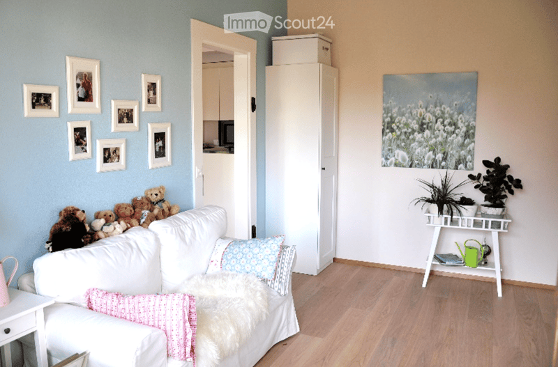 A cozy living room with a white sofa adorned with colorful pillows and a furry throw, surrounded by stuffed toy bears. A small table holds potted plants and a green watering can, with picture frames decorating the walls and a view into an adjacent kitchen