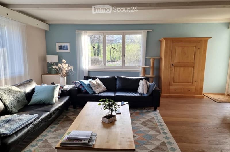Living room with light blue walls, wooden floor, large window with white curtains, black leather sofa with multiple cushions, wooden coffee table with a small plant, magazines, and a remote control, side table with a lamp, and a wooden cat tree.