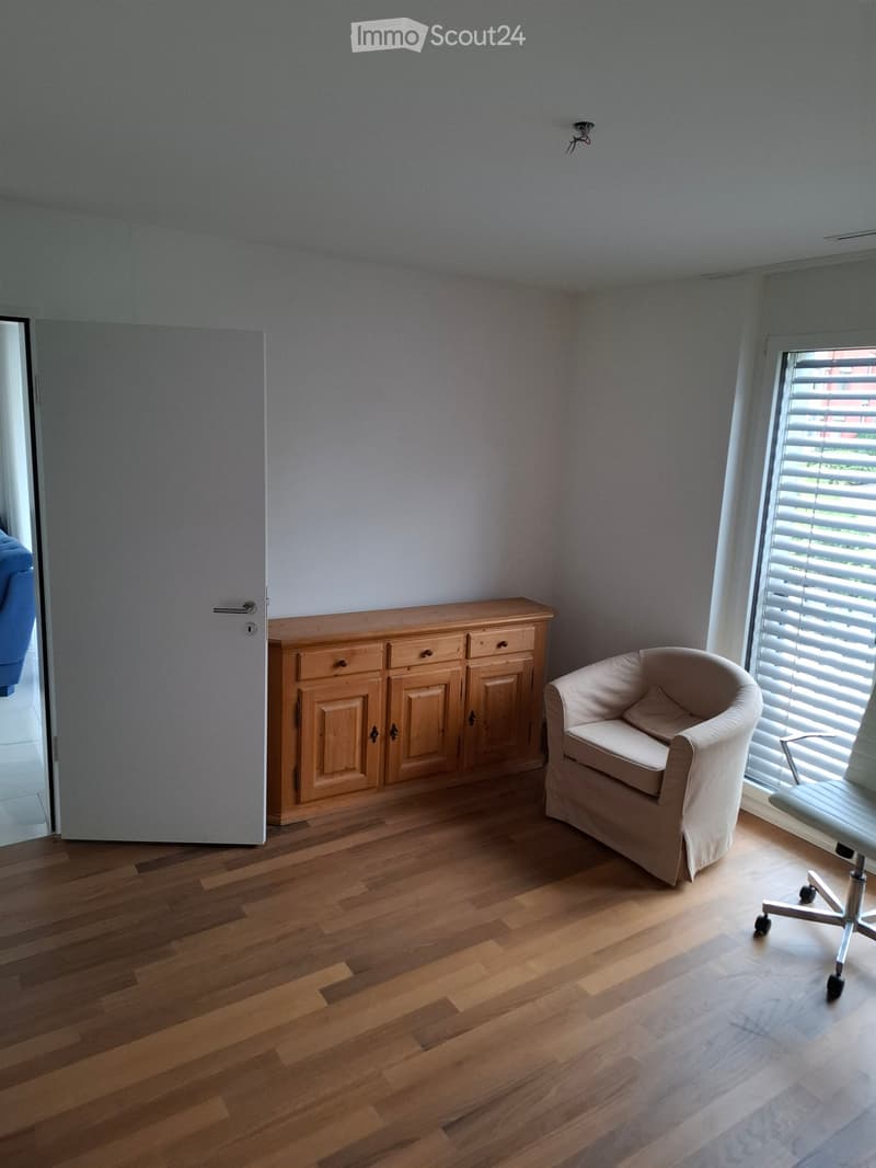 A furnished room with wooden flooring, a light-colored armchair, a wooden cabinet, and an office chair near a window with blinds.