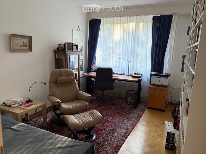 A well-furnished room with a large window, a comfortable leather chair with a matching footrest, a desk with a chair, a small table with books, a bookshelf, and a printer on a cabinet.