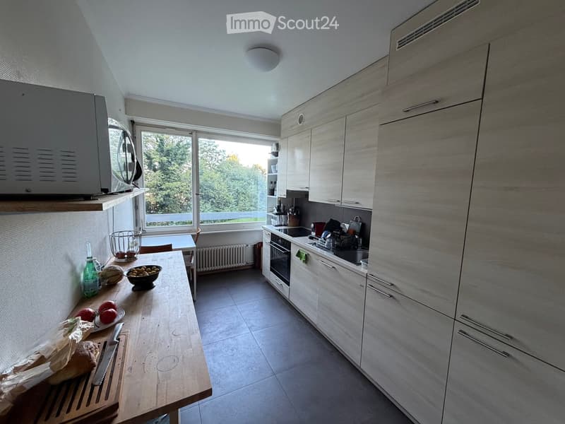 Modern kitchen with light wood cabinets, stainless steel appliances, and a large window overlooking greenery. The kitchen island has a wooden countertop with fruits, a bowl, and a bottle. The stove area includes an oven, microwave, and sink.
