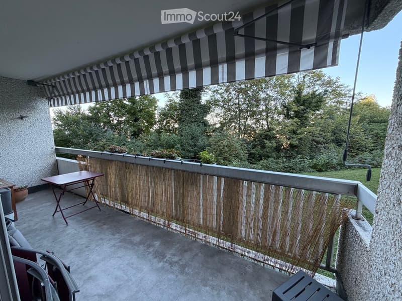 Balcony with a striped awning, bamboo privacy screen, small table, and chairs, overlooking a green area with trees.
