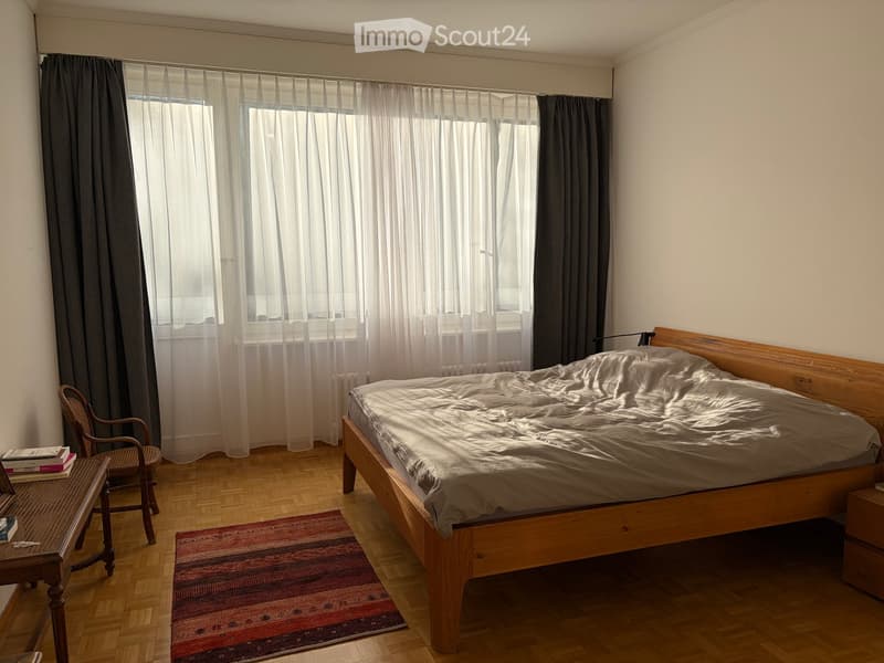 Bright bedroom with wooden bed, nightstand, wicker chair, small table with books, and a colorful rug on the floor.