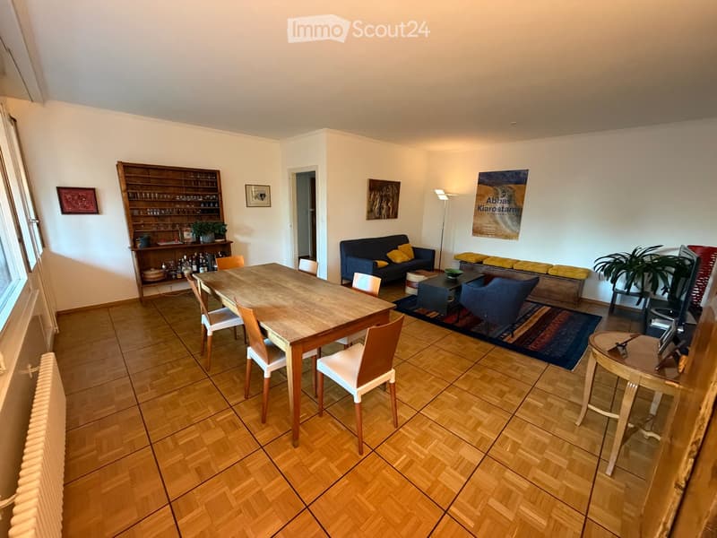 A well-lit living and dining area with wooden flooring, a sofa, a dining table with chairs, a cabinet, and a floor lamp.