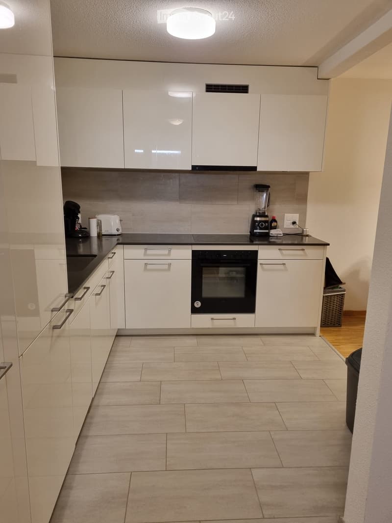 Modern kitchen with white cabinets, black countertops, and tiled floor. Appliances include an oven, blender, coffee maker, and toaster.